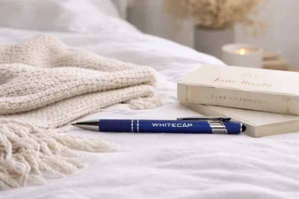 Blue engraved metal pen with WHITECAP logo on a bed beside books and a blanket