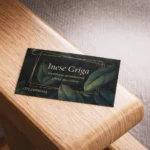 Vizītkarte uz koka virsmas ar tumšu lapu dizainu un zelta tekstu