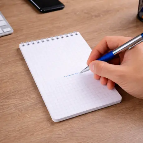 Hand writing with a blue pen in a spiral custom notepad on a wooden desk