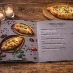 Atvērta A4 daudzlapu ēdienkarte ar hačapuri foto un cenrādi uz koka galda sveču gaismā