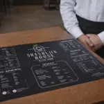 Melna A3 formāta vienlapas ēdienkarte uz koka galda restorānā ar viesmīli fonā