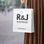 Balts papīra maisiņš ar R&J Boutique apdruku sievietes rokās