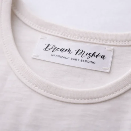 Gaišā apģērbā iešūta satīna birka ar melnu Dream Mishka druku